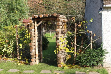 Pergola en rondins de bois entourée de végétation grimpante et d’un jardin paysager verdoyant.