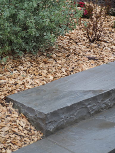 Marche en grès brut intégrée dans un massif avec paillage et arbustes décoratifs.