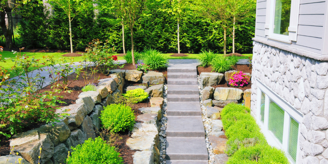 Aménagement extérieur paysager avec escalier en pierre, massifs fleuris et arbustes, autour d’une maison en pierre.