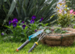 Outils de jardinage posés sur l’herbe près d’un panier de plantation, entourés de fleurs et de verdure.